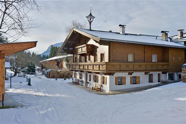 Een schilderachtig houten huis in de winter met een met sneeuw bedekte grond. Op de achtergrond zijn besneeuwde bergen en een heldere lucht te zien.