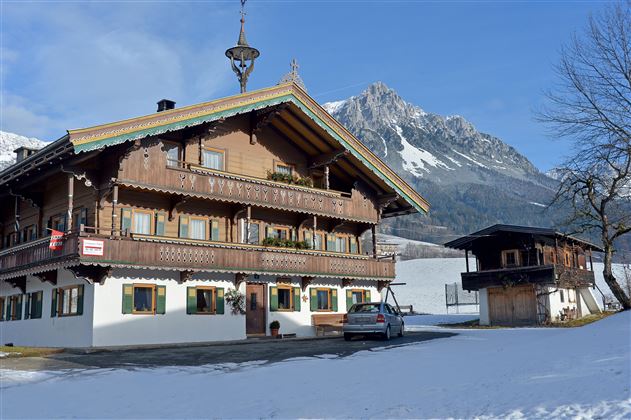 Ein traditionelles Holzhaus im Winter mit Schnee bedeckt. Im Hintergrund sind majestätische Berge und ein blauer Himmel zu sehen.