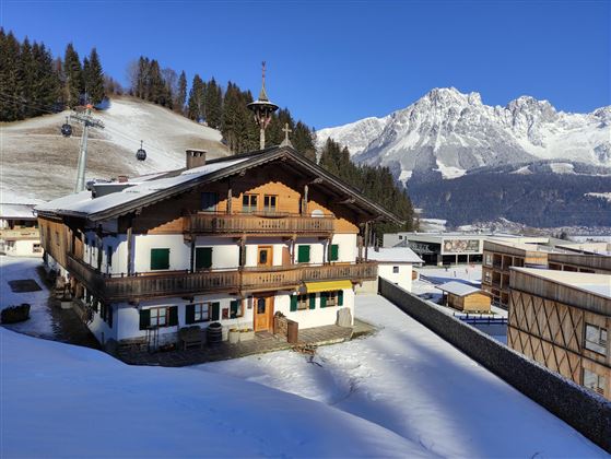 A traditional wooden house in a snowy landscape. In the background, there are high mountains and a cable car.