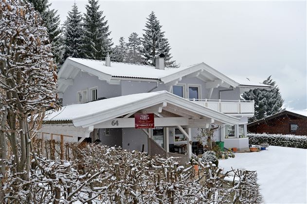 Ein charmantes Haus im Schnee, umgeben von verschneiten Bäumen. Der Himmel ist grau und bewölkt, was eine ruhige Winteratmosphäre schafft.