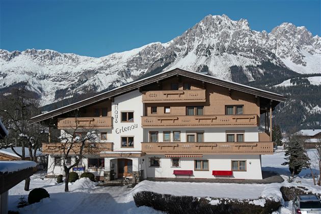Ein charmantes, mehrstöckiges Alpenhaus im Winter. Im Hintergrund erheben sich majestätische Berge, während der Boden mit Schnee bedeckt ist.