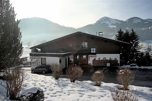 Ein charmantes Haus im Schnee, umgeben von Bergen und Bäumen. Die Sonne scheint, was eine warme Atmosphäre schafft.