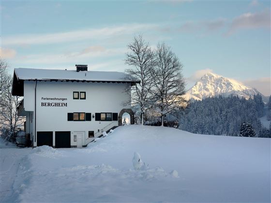 A picturesque holiday home in the snow named "Bergheim". In the background, snow-covered mountains and a clear sky can be seen.