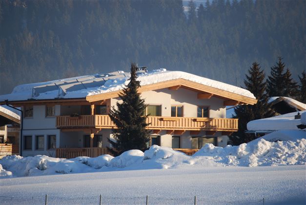 A cozy house in the snow with a large balcony. In the background, snow-covered trees and mountains can be seen.