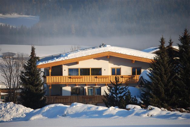 A cozy house with a wooden balcony, surrounded by snow-covered trees. The landscape is picturesque and wintry.