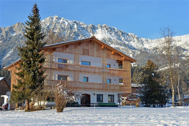 Ein charmantes Holzhaus in einer verschneiten Landschaft, umgeben von majestätischen Bergen. Die klare Winterluft verleiht der Szene eine ruhige Atmosphäre.