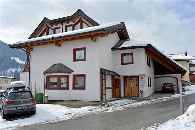 A modern house in the mountains, surrounded by snow. The facade is white with wooden details and large windows.