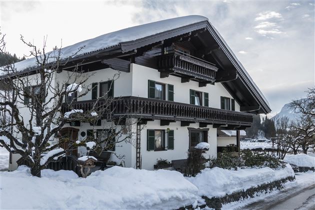 Ein traditionelles Haus im bayerischen Stil, umgeben von Schnee. Der Himmel ist bewölkt und die Umgebung wirkt ruhig und malerisch.