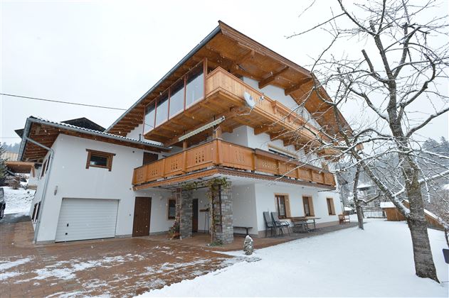 Ein modernes Haus im Schnee mit einem großen Balkon aus Holz. Die Umgebung ist ruhig und malerisch, ideal für einen Winteraufenthalt.