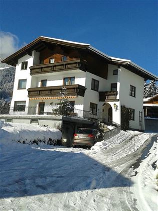 A modern house in the snow with wooden-clad balconies. The parking lot is covered with snow and the surroundings show wintery mountains.