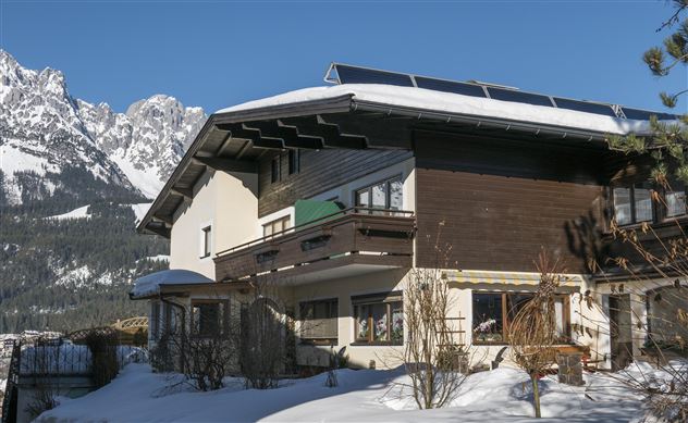 Ein gemütliches Haus im Schnee, umgeben von beeindruckenden Bergen. Der klare blaue Himmel ergänzt die winterliche Landschaft.
