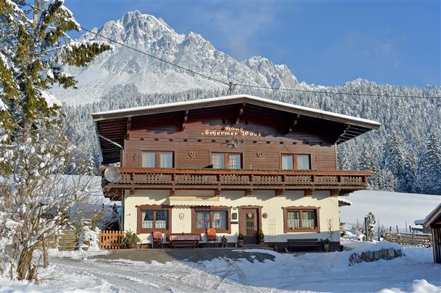 A charming wooden house in winter, surrounded by snow-covered trees and mountains. The clear blue sky shines over the peaceful landscape.