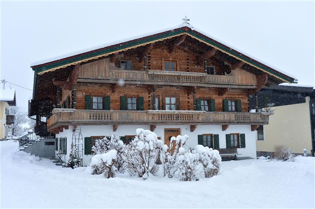 A traditional Alpine house with a wooden structure, surrounded by snowy terrain. The sky is gray and there are snowdrifts in front of the building.
