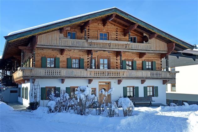 A traditional wooden house with several balconies and green shutters. The entire scene is surrounded by snow and exudes a wintry atmosphere.