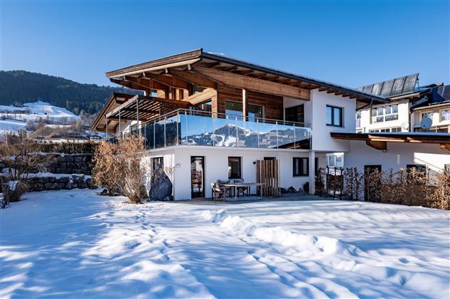 Ein modernes Haus mit Holzveranda im Schnee. Im Hintergrund sind schneebedeckte Berge zu sehen und der Himmel ist klar und blau.