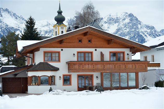 Ein traditionelles Haus im alpinen Stil, umgeben von Schnee. Im Hintergrund sind beeindruckende Berge zu sehen.