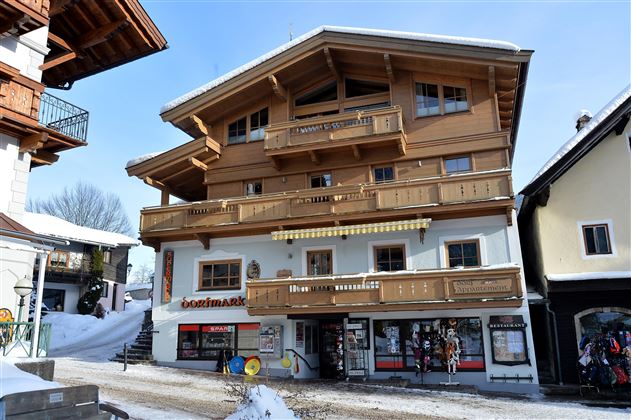 Ein traditionelles Holzhaus in den Alpen mit mehreren Balkonen. Vor dem Gebäude sind Geschäfte und Schnee zu sehen.