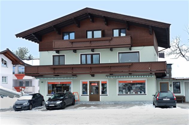 A two-story building in alpine style with wooden cladding and balconies. In front of the house, several cars are parked in a snow-covered area.