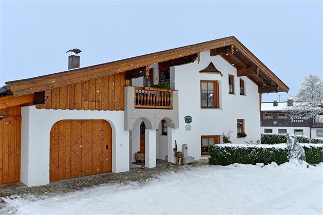 Ein charmantes Haus im alpinen Stil, umgeben von Schnee. Der Dachstuhl ist aus Holz, und es gibt eine kleine Terrasse mit Pflanzen.