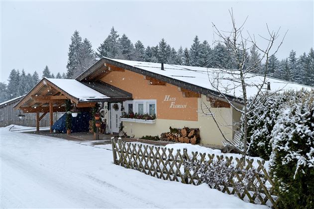 Ein gemütliches Haus in einer verschneiten Landschaft. Bäume im Hintergrund und ein weißer Zaun ergänzen die winterliche Atmosphäre.
