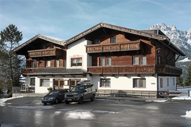 A typical alpine house with a wooden facade and balconies. In front of the house are several cars, surrounded by snow-covered ground.