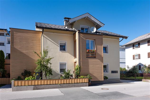 Ein modernes Gebäude mit einer Mischung aus Holz- und Putzfassade. Es liegt an einer ruhigen Straße und hat einen klaren blauen Himmel im Hintergrund.