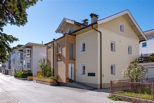 Ein modernes Haus an einer ruhigen Straße mit klar blauem Himmel. Die Gebäude sind von Bäumen und grünen Pflanzen umgeben.