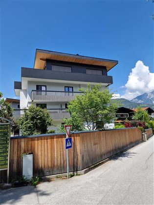 Ein modernes Haus mit mehreren Etagen und einer Dachterrasse. Umgeben von Bäumen und einem gepflegten Garten in einer ruhigen Straße.