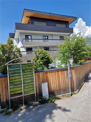 Ein modernes Gebäude mit mehreren Balkonen und einer begrünten Umgebung. Auf der linken Seite ist ein Informationsschild mit verschiedenen Angeboten zu sehen.