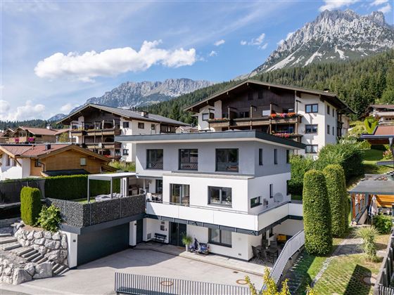 Ein modernes Haus mit zwei Etagen in einer malerischen Berglandschaft. Im Hintergrund sind hohe Berge und weitere Wohnhäuser sichtbar.