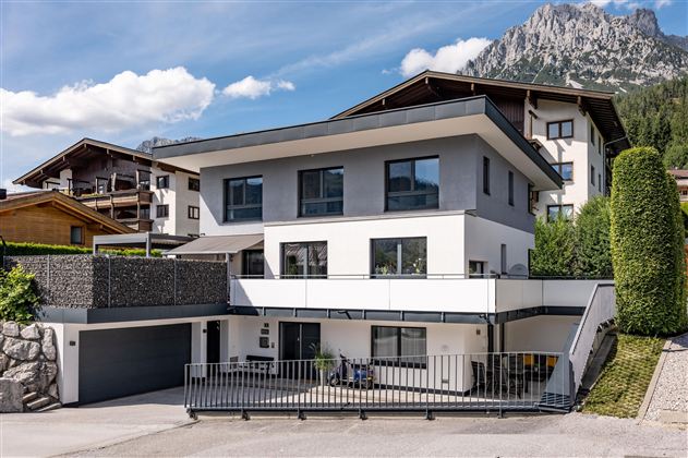 Ein modernes Haus mit großen Fenstern und einem klaren Parkplatz. Im Hintergrund sind Berge und eine schöne Landschaft sichtbar.