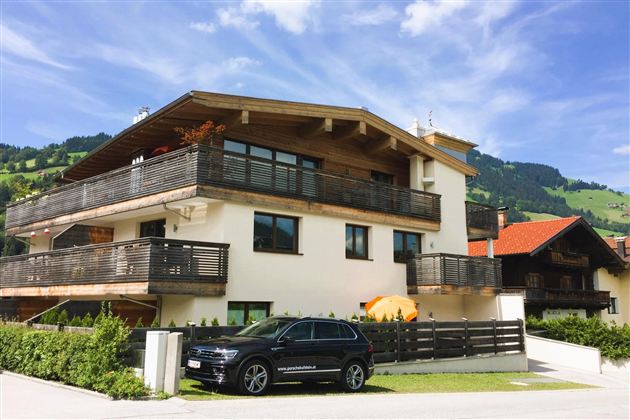 Ein modernes Haus mit mehreren Balkonen und großen Fenstern. Im Vordergrund steht ein schwarzes Auto und im Hintergrund sind grüne Berge zu sehen.