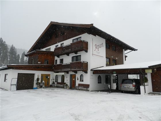 Ein gemütliches Gebäude im alpinen Stil, umgeben von Schnee. Die Fassade ist mit Balkonen und einem Eingang ausgestattet, der in eine winterliche Landschaft führt.