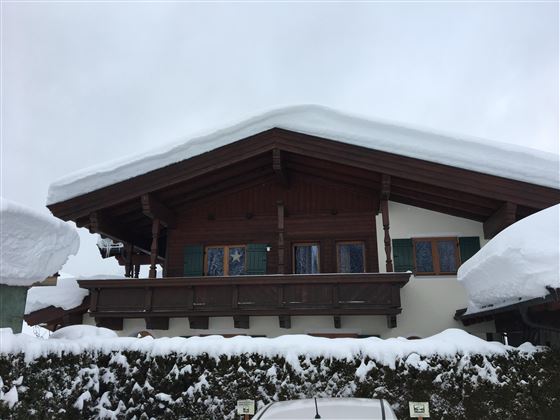 A chalet-style house with a snow-covered roof and large windows. The area in front of the house is also covered with snow.