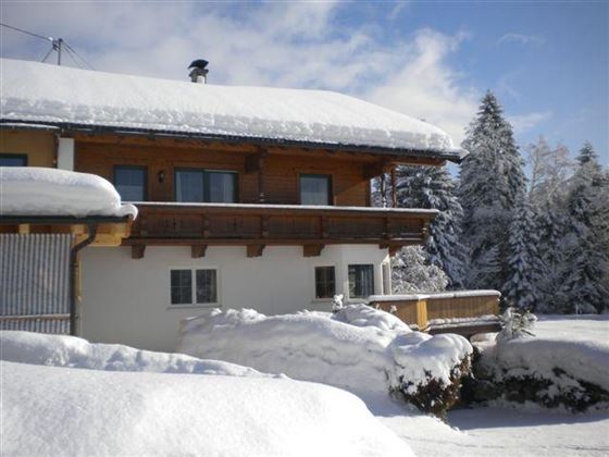 A cozy house in the snow with a wooden balcony. All around, the landscape is surrounded by snow-covered trees.