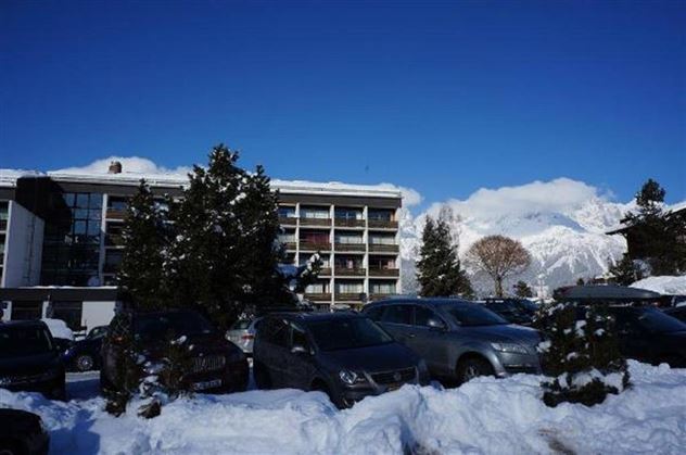 Ein Gebäude, umgeben von Schnee, mit einem klaren blauen Himmel im Hintergrund. Parkende Autos und Bäume sind ebenfalls sichtbar.
