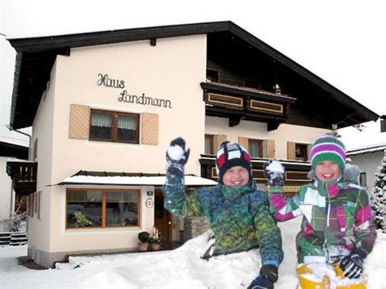 Zwei fröhliche Kinder spielen im Schnee vor einem gemütlichen Haus. Das Haus trägt die Aufschrift "Haus Landmann" und ist von Schnee umgeben.