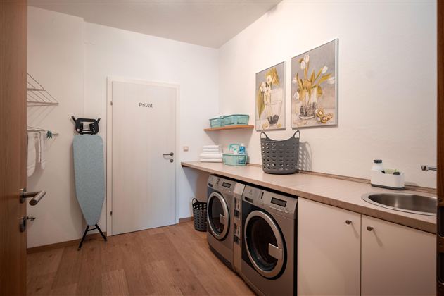 Ein moderner Waschraum mit zwei Waschmaschinen und einem Wäschetrockner. An der Wand hängen Bilder und es stehen verschiedene Aufbewahrungskörbe.