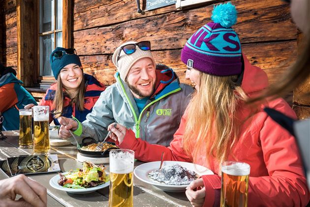 Eine Gruppe von Freunden sitzt an einem Tisch in einer Hütte. Sie genießen Essen und Bier und lachen zusammen.