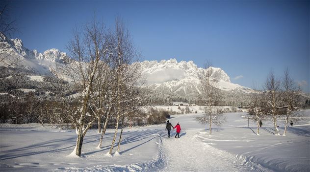 Eine verschneite Landschaft mit hohen Bergen im Hintergrund. Zwei Personen gehen einen Weg, umgeben von schneebedeckten Bäumen.
