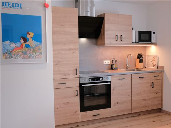 A modern kitchen with wooden cabinets, a stove, and a microwave. A colorful picture of "Heidi" hangs on the wall.