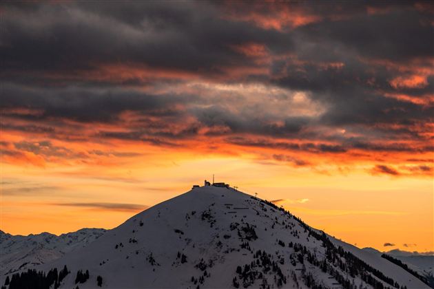Ein schneebedeckter Berg unter einem orangefarbenen Sonnenuntergang. Die Wolken sind dramatisch und verleihen der Szene eine mystische Stimmung.