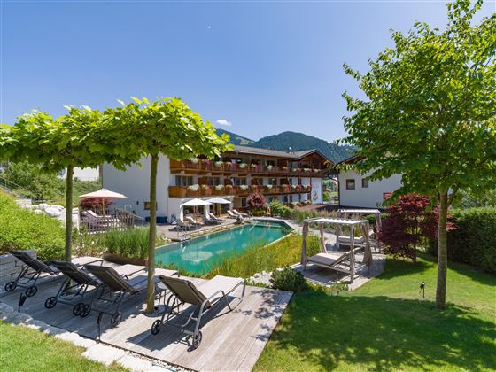 Außenansicht des Hotels Alpenpanorama mit Garten, Pool, Sonnenliegen und Blick auf die Tiroler Bergwelt.