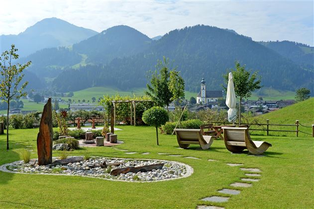 Gartenanlage des Hotel Alpenpanorama in der Ferienregion Wilder Kaiser mit Sonnenliegen, Rundbank und Panoramablick auf die Tiroler Berge.