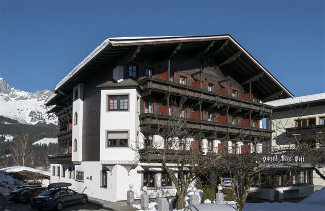 Ein traditionelles Alpenhotel mit mehreren Balkonen und einem großen Dach. Schnee liegt rund um das Gebäude, und die Berge sind im Hintergrund sichtbar.