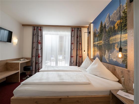 Ein gemütliches Zimmer mit zwei Betten und Holzdekor. An der Wand befindet sich ein großes Landschaftsbild und es gibt viel Tageslicht durch die Fenster.