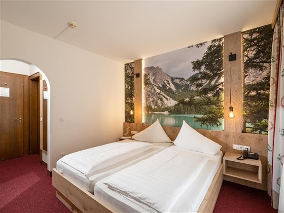 Ein gemütliches Schlafzimmer mit einem großen Bett und einem schönen Wandbild von Bergen an einem See. Der Raum hat eine warme Atmosphäre mit sanfter Beleuchtung.