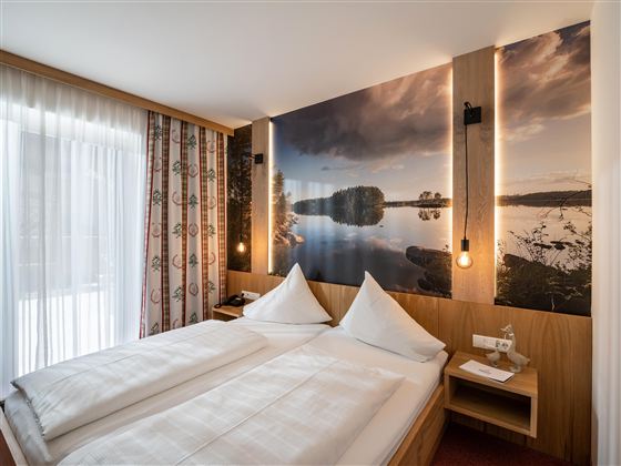 Ein modernes Schlafzimmer mit zwei Betten und einer schönen Fototapete, die eine Landschaft am Wasser zeigt. Große Fenster sorgen für viel Tageslicht.