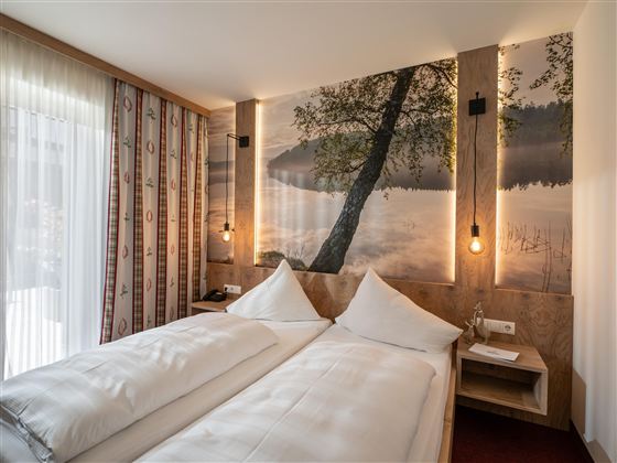 Ein modernes Hotelzimmer mit zwei Einzelbetten und einem stilvollen Wandmotiv eines Sees. Sanfte Beleuchtung und große Fenster sorgen für eine helle, einladende Atmosphäre.