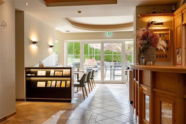 Ein einladender Empfangsbereich mit hellen Wänden und einer modernen Theke. Große Fenster bieten einen Blick auf eine Terrasse mit Sitzplätzen im Freien.